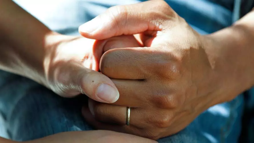 Two hands gently clasp each other, conveying support and compassion. One hand wears a simple ring.