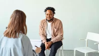 A man is sitting on a chair, talking to a woman holding a clipboard with her back to the camera