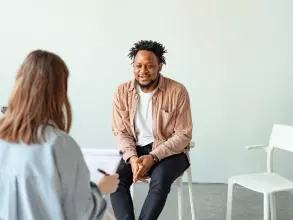 A man is sitting on a chair, talking to a woman holding a clipboard with her back to the camera